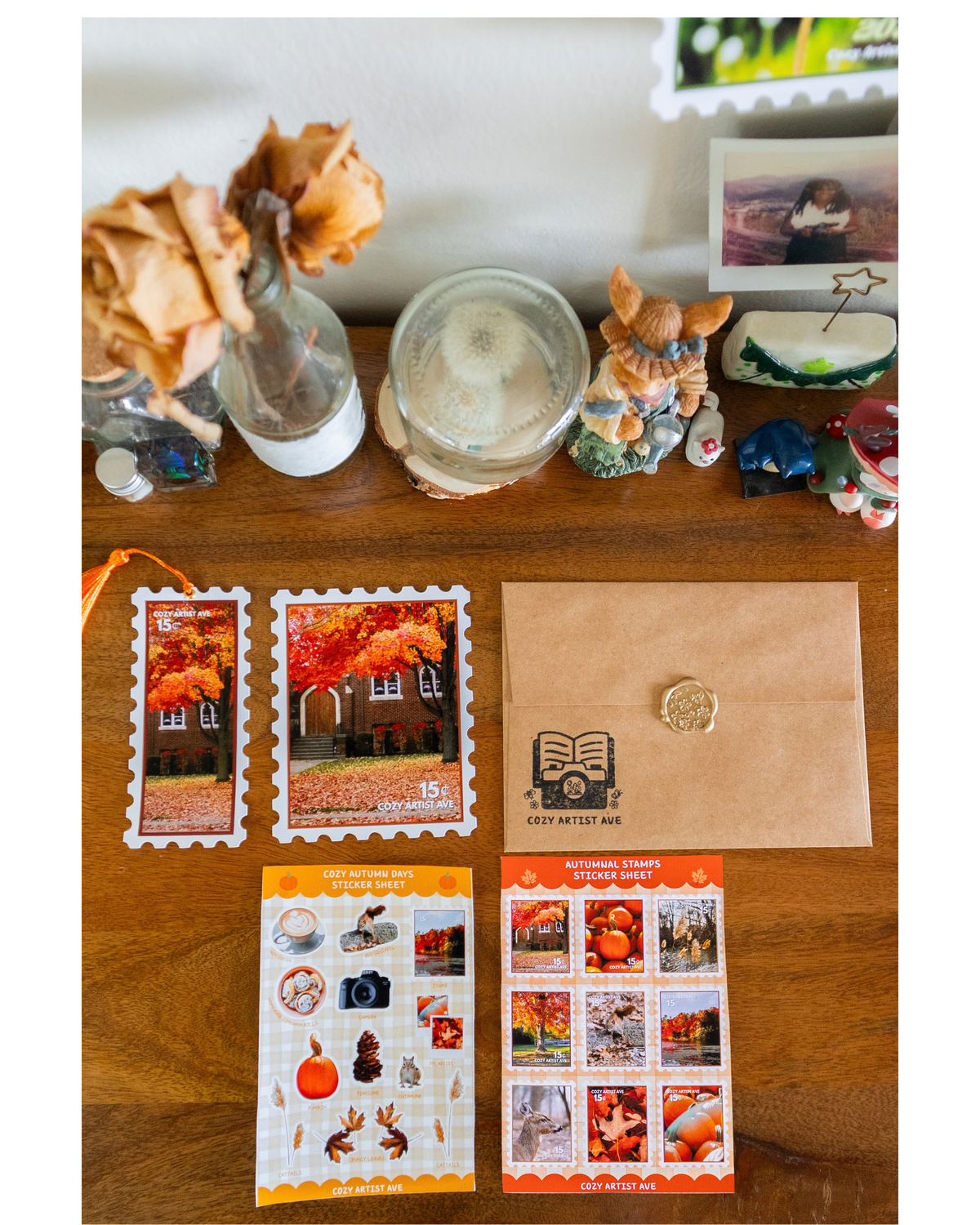 A styled flat-lay of autumn-themed postage stamps and a brown envelope on a wooden tabletop, surrounded by decorative home items like dried flowers, a candle, and small figurines.